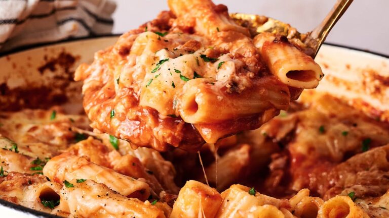A close-up of baked ziti being served from a dish, showing pasta, tomato sauce, melted cheese, and herbs.