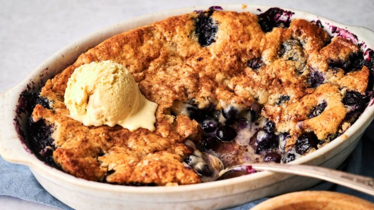 A baked blueberry cobbler with a golden crust, partially served, topped with a scoop of vanilla ice cream.