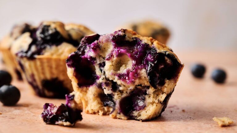 A close-up of a blueberry muffin with a bite taken out, showing the moist interior and whole blueberries. Other muffins and blueberries are in the background.