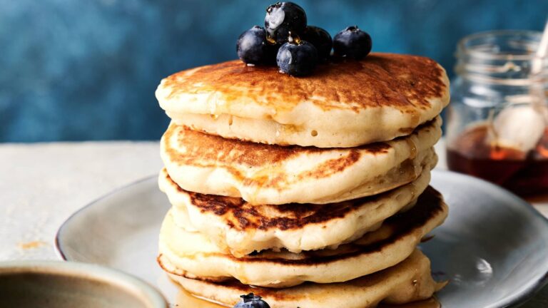 A stack of golden-brown pancakes topped with blueberries and syrup sits on a plate with a jar of syrup in the background.