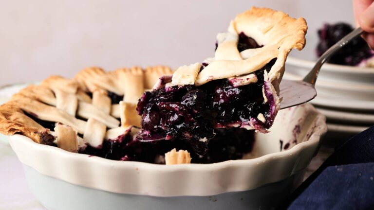 A slice of blueberry pie with a lattice crust is being lifted from a white ceramic pie dish.