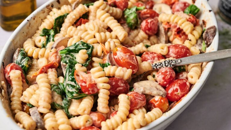 A white oval dish filled with pasta, cherry tomatoes, spinach, and mushrooms in a creamy sauce, with a metal serving spoon.