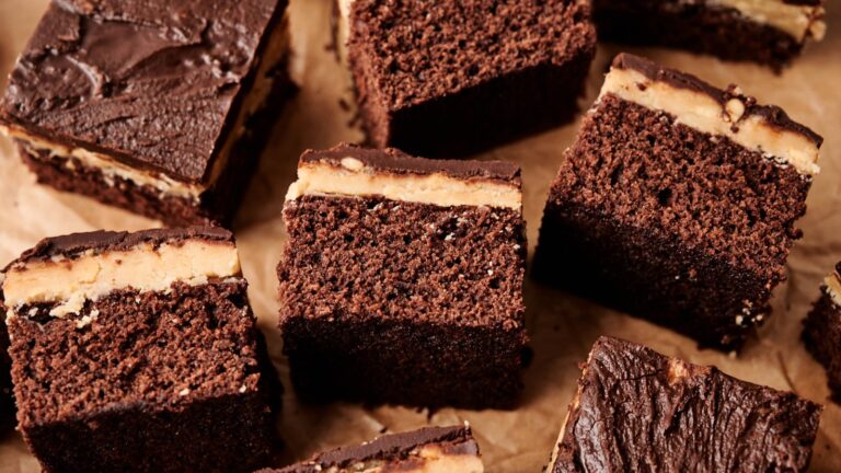 Several chocolate brownie squares with a peanut butter layer and chocolate frosting are arranged on a sheet of parchment paper.