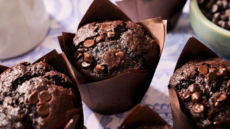 Four chocolate muffins with chocolate chips sit in brown paper wrappers on a patterned surface.