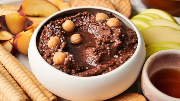 A white bowl of chocolate hummus topped with chickpeas and coarse salt, surrounded by sliced peaches, apples, cookies, and rolled wafers.