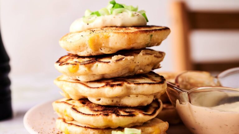 A stack of savory pancakes topped with a dollop of sauce and chopped green onions, with a small bowl of dipping sauce beside them on a plate.
