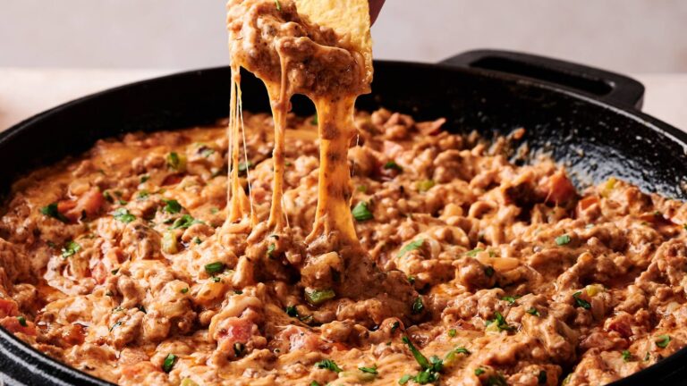 A hand dips a tortilla chip into a skillet filled with cheesy, ground beef queso dip mixed with tomatoes and herbs.