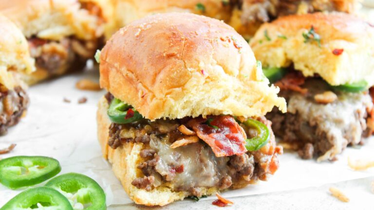 A close-up of a sandwich slider filled with ground beef, melted cheese, bacon, and jalapeño slices on a soft bun, with more sliders and jalapeño slices in the background.