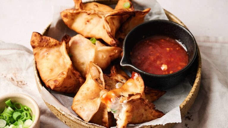 A bowl of fried wontons filled with cream cheese, served with a side of sweet chili dipping sauce and garnished with chopped green onions.