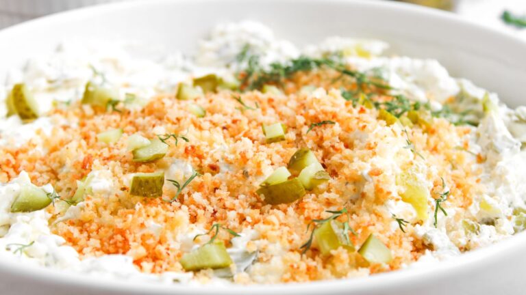 A close-up of a creamy dip topped with golden breadcrumbs, chopped pickles, and fresh dill in a white bowl.