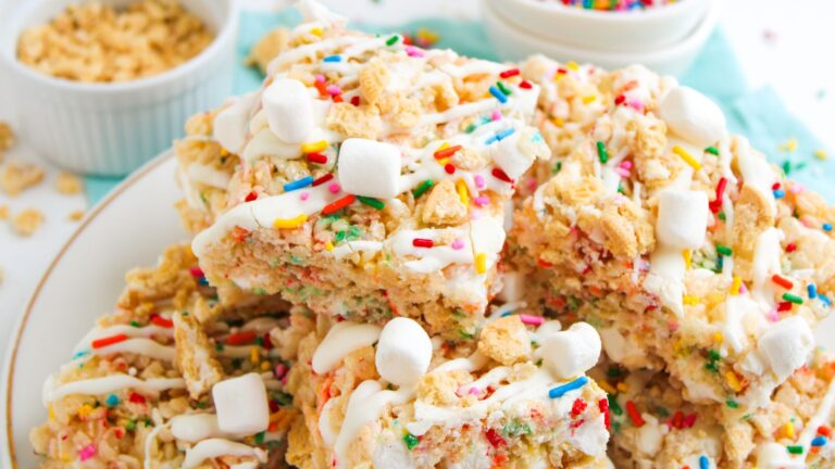 A plate of cereal marshmallow bars topped with mini marshmallows, rainbow sprinkles, and a white icing drizzle, with bowls of cereal and sprinkles in the background.