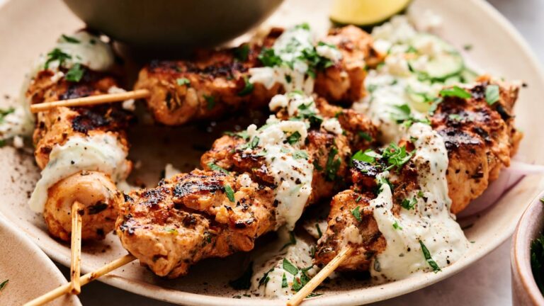 Grilled chicken skewers topped with a creamy white sauce and chopped herbs, served on a plate with sliced vegetables.