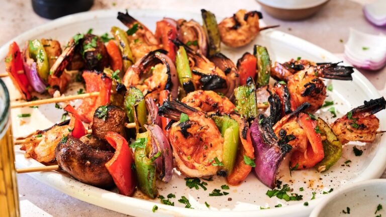 A white plate holds grilled shrimp and vegetable skewers with mushrooms, bell peppers, onions, and tomatoes, garnished with chopped herbs.