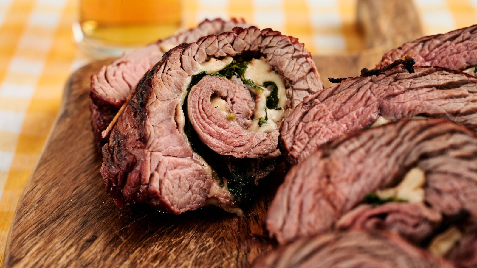 Close-up of sliced stuffed beef roulade with visible layers of meat, cheese, and spinach on a wooden board. A glass of beer is blurred in the background.