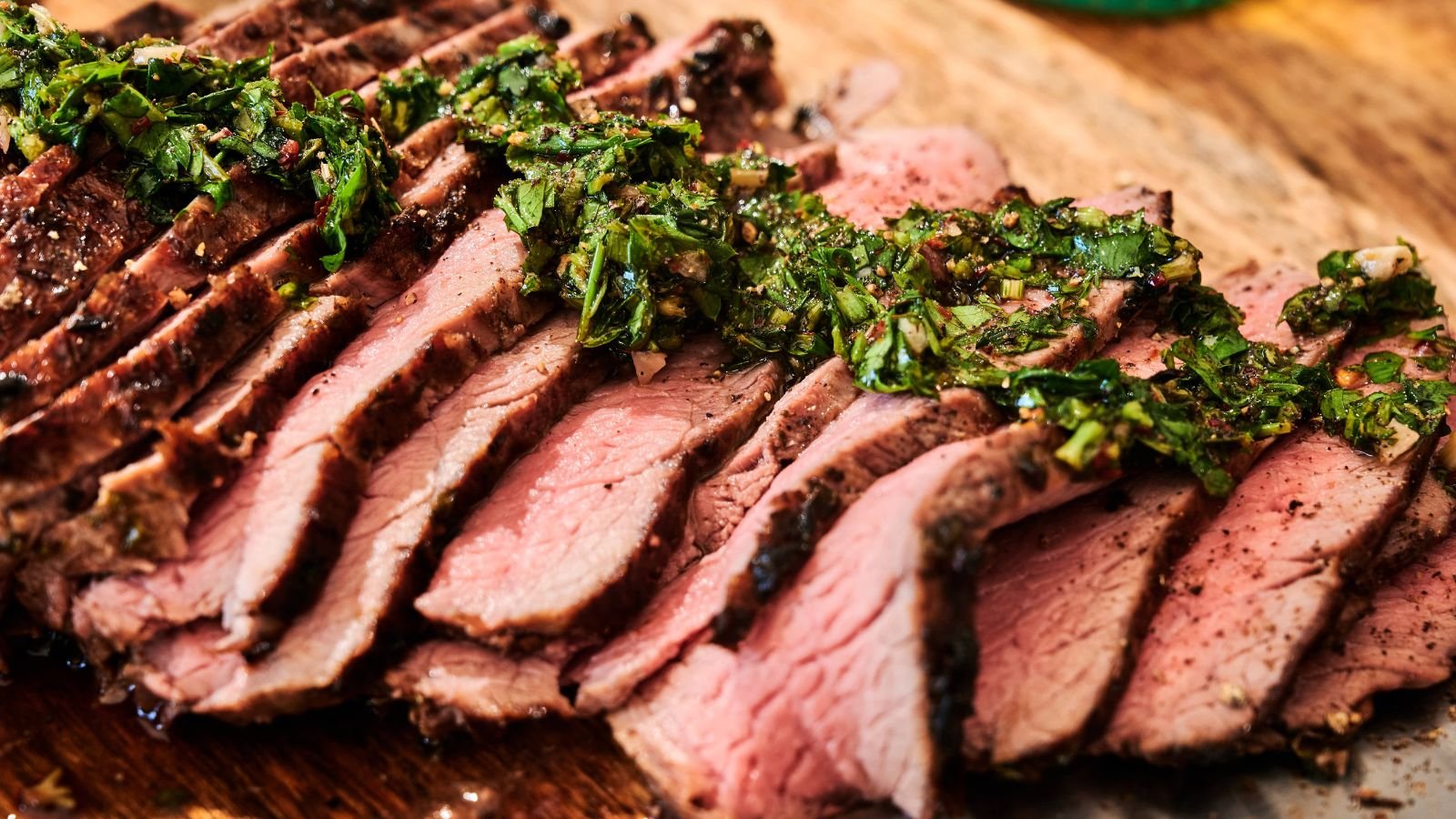 Sliced medium-rare steak arranged on a wooden board, topped with a green herb sauce.