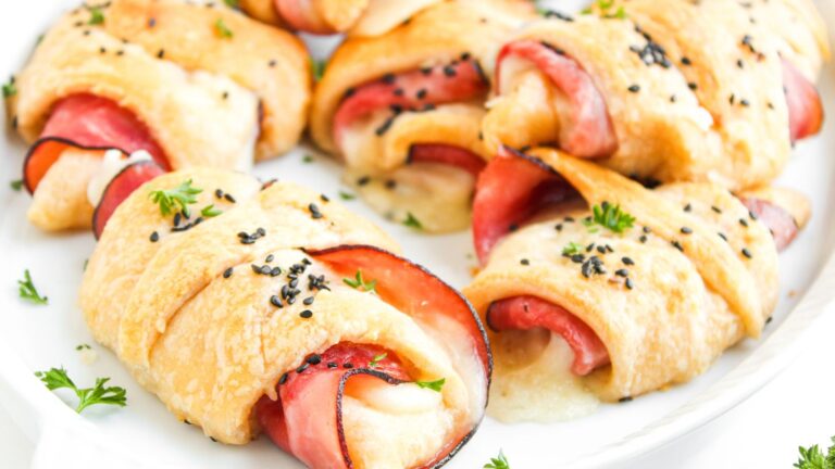 Crescent rolls filled with ham and melted cheese, topped with black sesame seeds and garnished with chopped parsley on a white plate.
