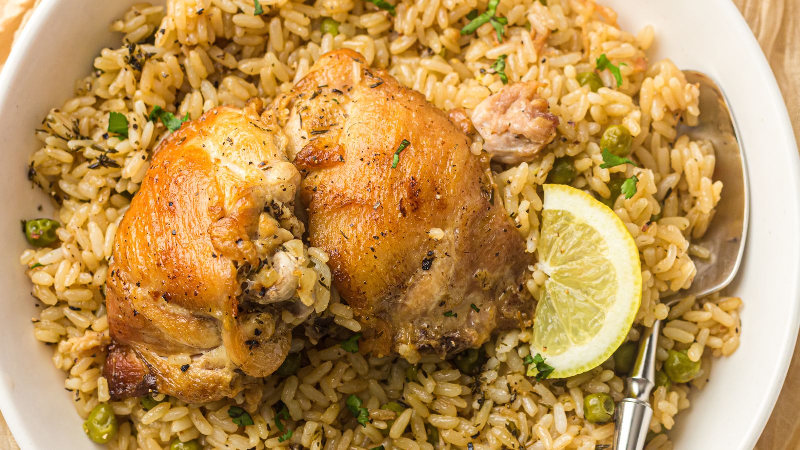 A plate of seasoned rice with green peas, topped with two pieces of roasted chicken and a lemon slice, served with a spoon.