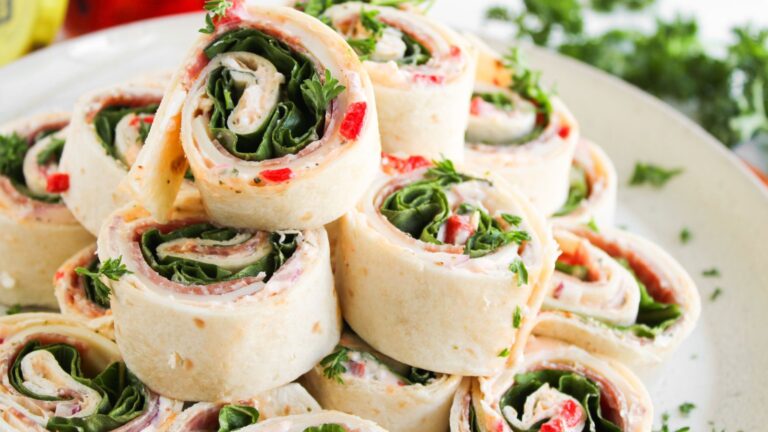 A plate of tortilla pinwheel sandwiches filled with spinach, cream cheese, red peppers, and herbs, stacked neatly on top of each other.