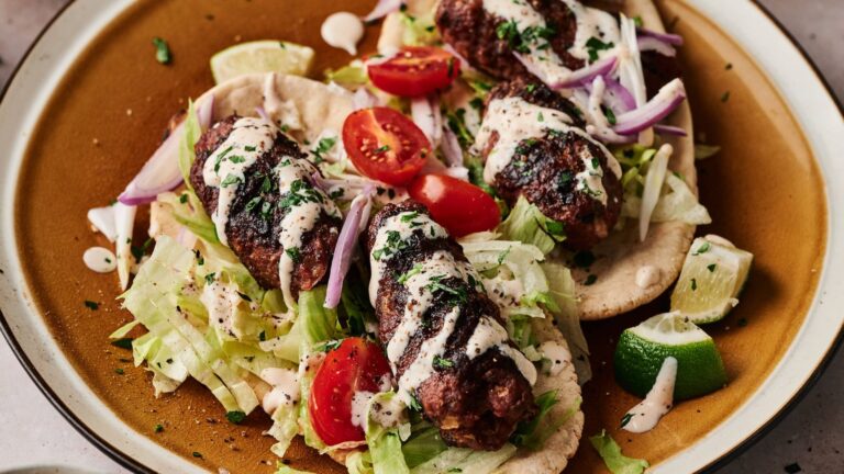 A plate with grilled meat skewers on flatbread, topped with lettuce, cherry tomatoes, red onion, herbs, lime wedges, and a drizzle of creamy sauce.