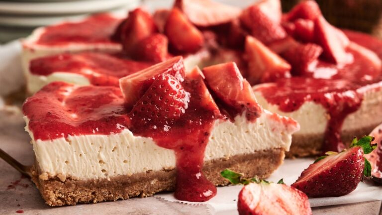 A slice of cheesecake with a crumbly base, topped with strawberry sauce and fresh strawberry pieces, is being served.