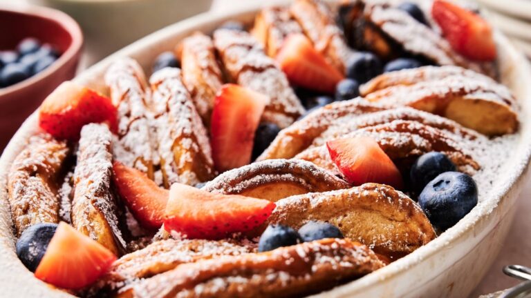 A baked French toast casserole topped with powdered sugar, fresh strawberries, and blueberries in a white dish.