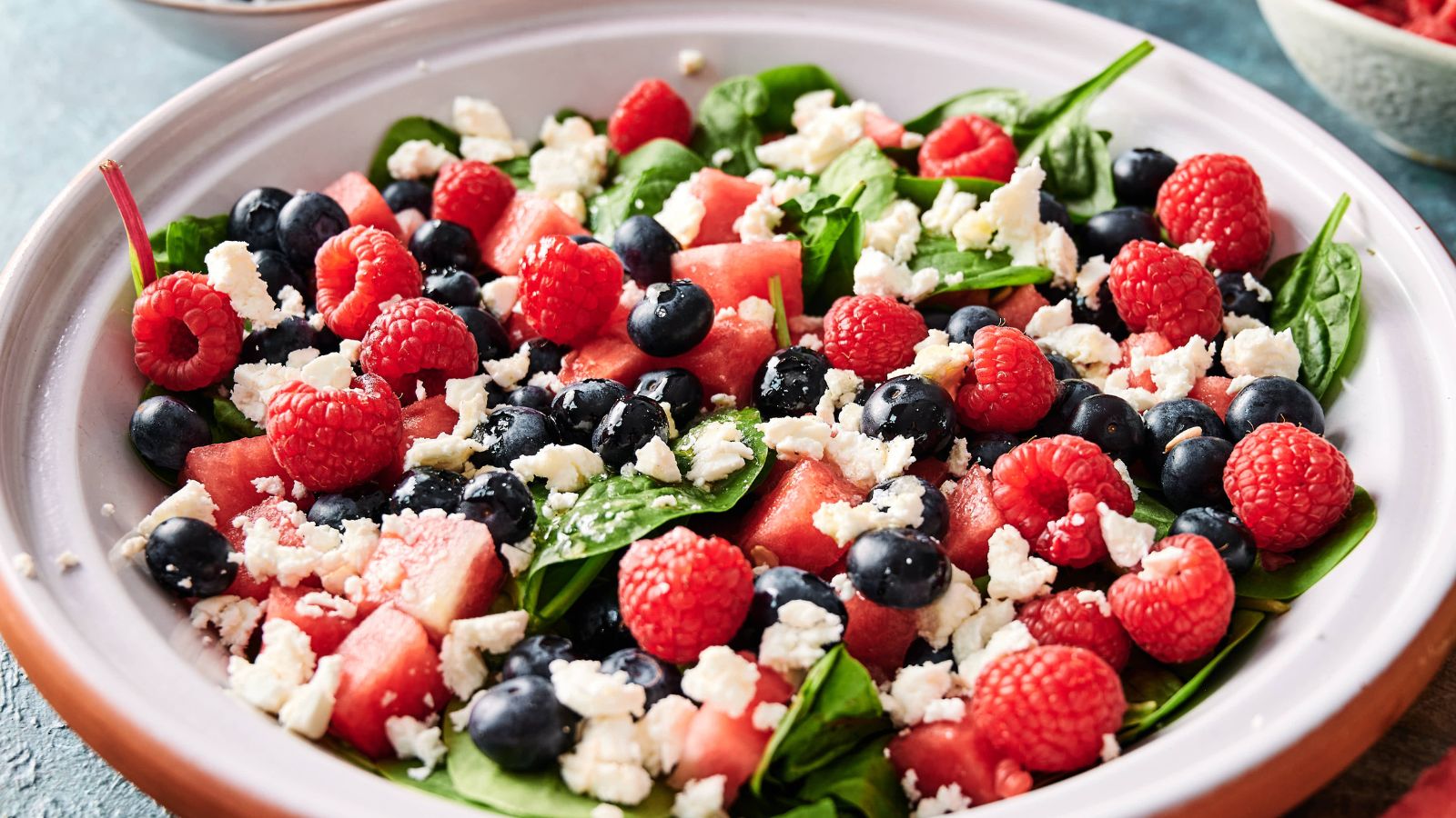 A salad with spinach leaves, watermelon cubes, blueberries, raspberries, and crumbled feta cheese in a white bowl.