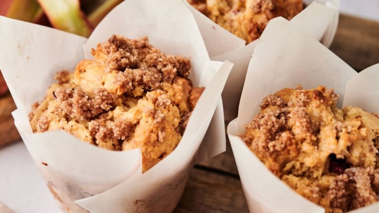 Close-up of three muffins with crumbly streusel topping, each wrapped in white parchment paper, arranged on a wooden surface.