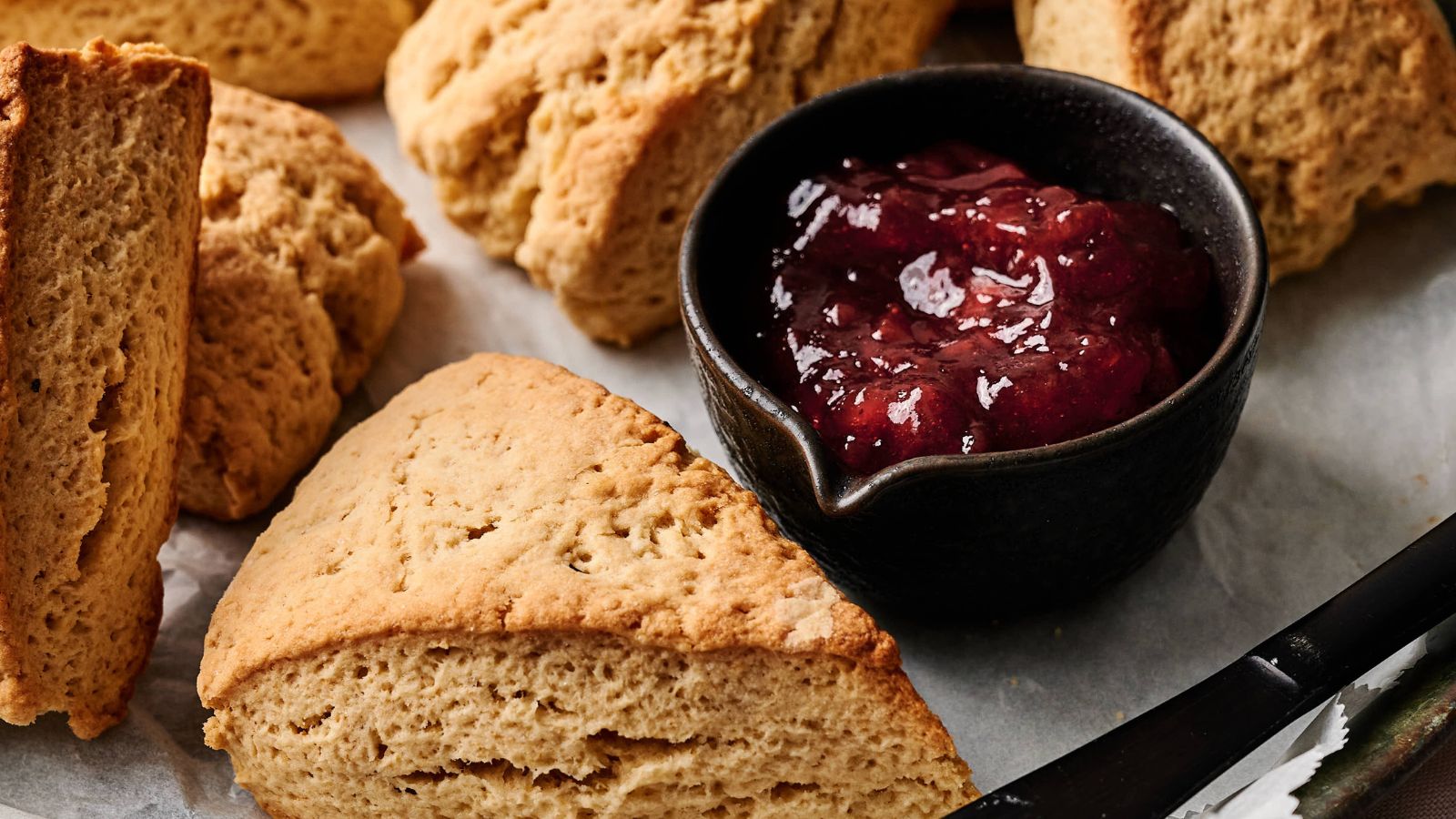 Triangular scones arranged on a tray with a small black bowl of red fruit jam placed in the center.