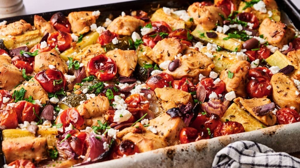 A baking tray filled with roasted chicken pieces, cherry tomatoes, zucchini, olives, and crumbled feta cheese, garnished with fresh herbs.
