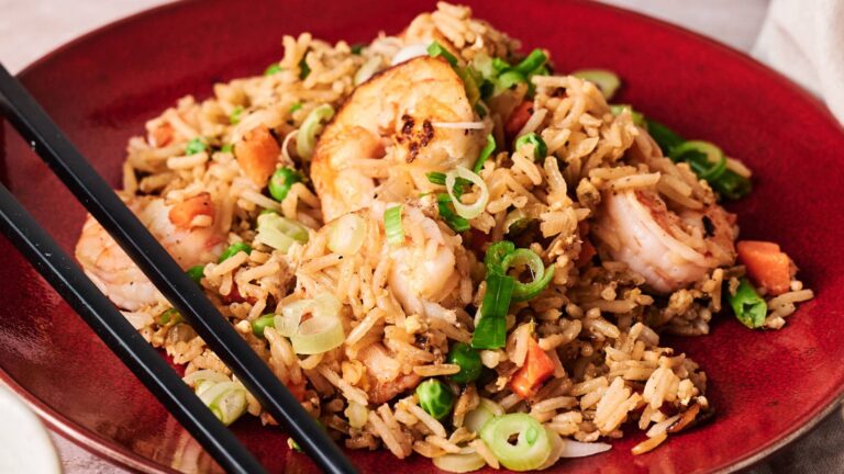 A plate of shrimp fried rice with chopped green onions and vegetables, served on a red dish with black chopsticks on the side.