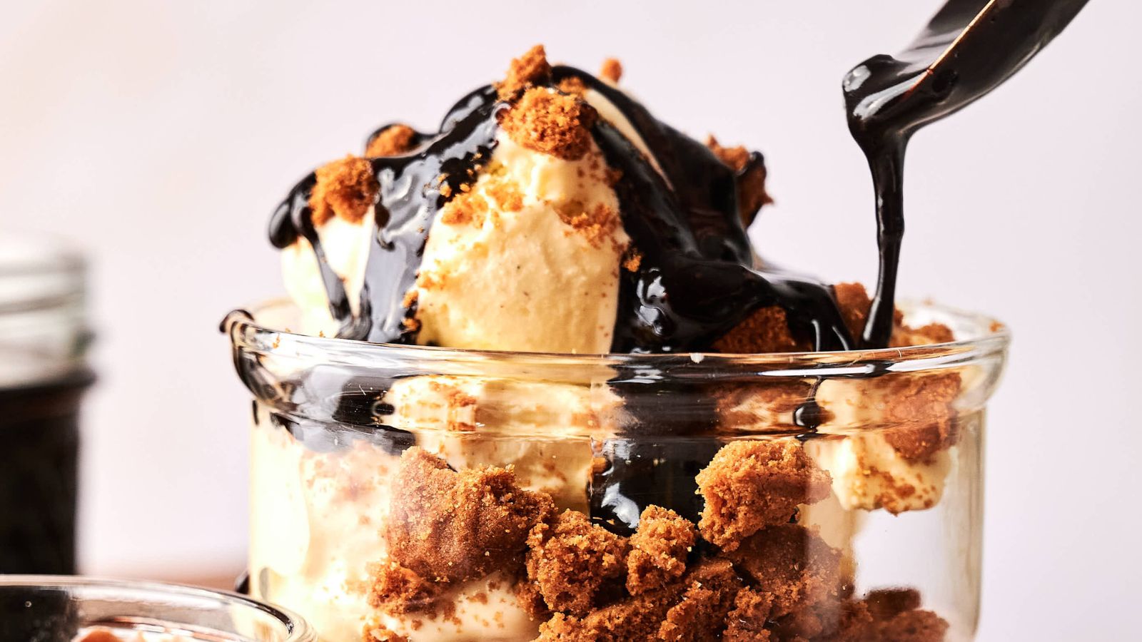 A glass filled with vanilla ice cream, crumbled cookies, and chocolate sauce being poured on top.