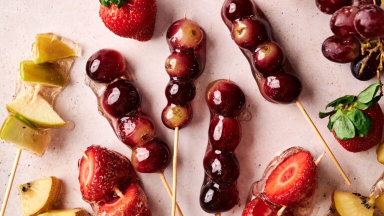 Assorted skewered fruits, including grapes, strawberries, and kiwi, coated in a glossy, clear candy shell, arranged on a light surface.