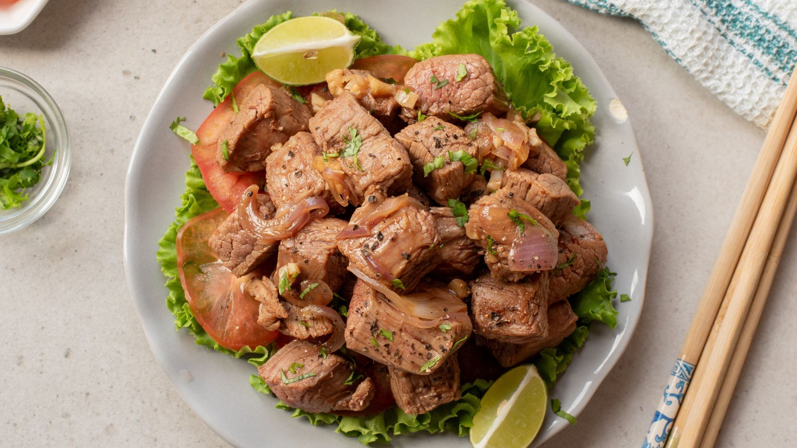 A plate of stir-fried beef cubes with onions, garnished with cilantro, served on lettuce with tomato slices and lime wedges. Chopsticks rest beside the plate.