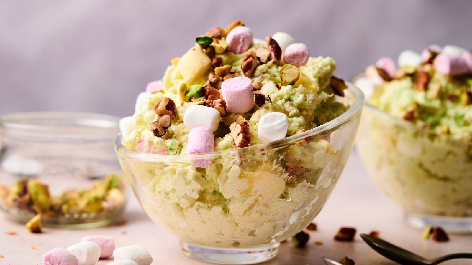 A glass bowl filled with pistachio-flavored dessert topped with mini marshmallows and chopped nuts, with another similar bowl in the background.