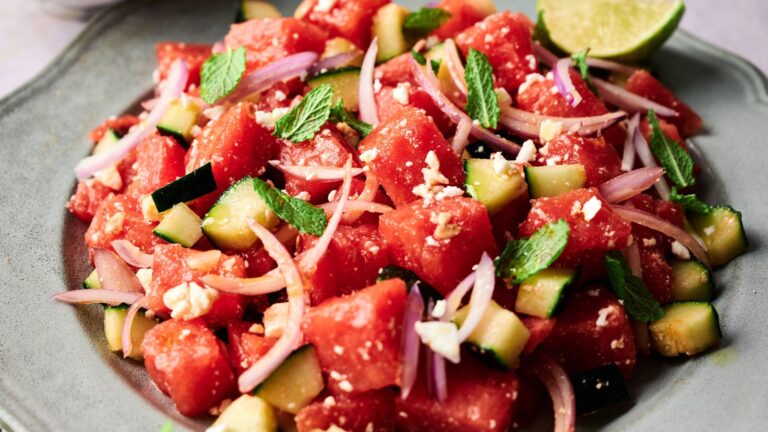 A plate of watermelon salad with cucumber, red onion, feta cheese, and mint leaves, garnished with a lime wedge.