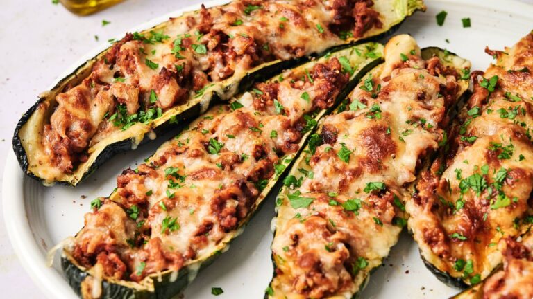 Four zucchini boats topped with ground meat, tomato sauce, melted cheese, and chopped parsley on a white plate.