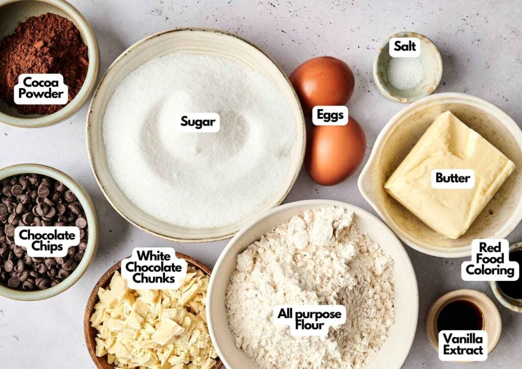 Assorted baking ingredients for red velvet brownies, including cocoa powder, chocolate chips, white chocolate chunks, sugar, flour, eggs, butter, salt, vanilla extract, and red food coloring arranged on a countertop.