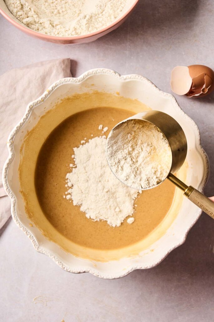 A measuring cup pours flour into a bowl of brown batter for rhubarb muffins, with a cracked eggshell and a bowl of flour nearby.
