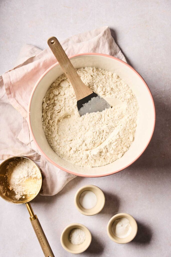 A mixing bowl with flour and a spatula sits on a cloth, surrounded by measuring cups containing flour, salt, and baking powder—ingredients ready to whip up delicious rhubarb muffins.