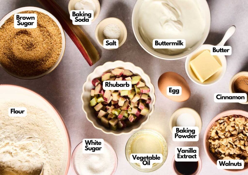 Various baking ingredients for Rhubarb Muffins, including flour, brown sugar, white sugar, rhubarb, egg, buttermilk, butter, walnuts, and labeled seasonings and leaveners are neatly arranged on a countertop.