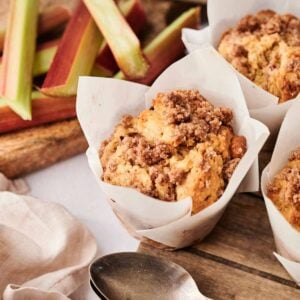 Three crumb-topped rhubarb muffins in parchment wrappers sit on a wooden surface, with fresh rhubarb stalks and a knife in the background, and two metal spoons in the foreground.