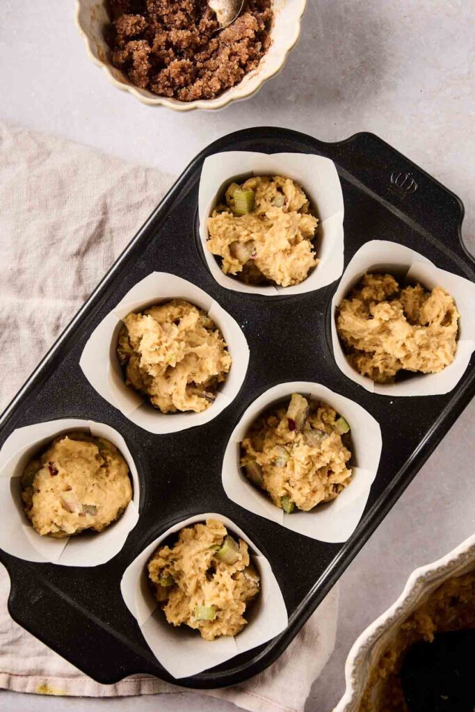 A muffin tin filled with six paper liners, each holding unbaked rhubarb muffins batter, sits on a table beside a bowl of brown sugar topping.