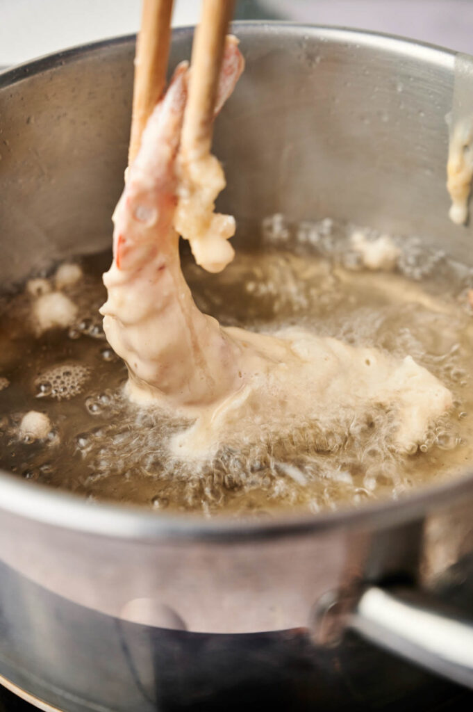 A breaded chicken wing is being lifted out of hot oil in a saucepan with chopsticks while deep frying, reminiscent of the crispy texture found in a Shrimp Tempura Roll.