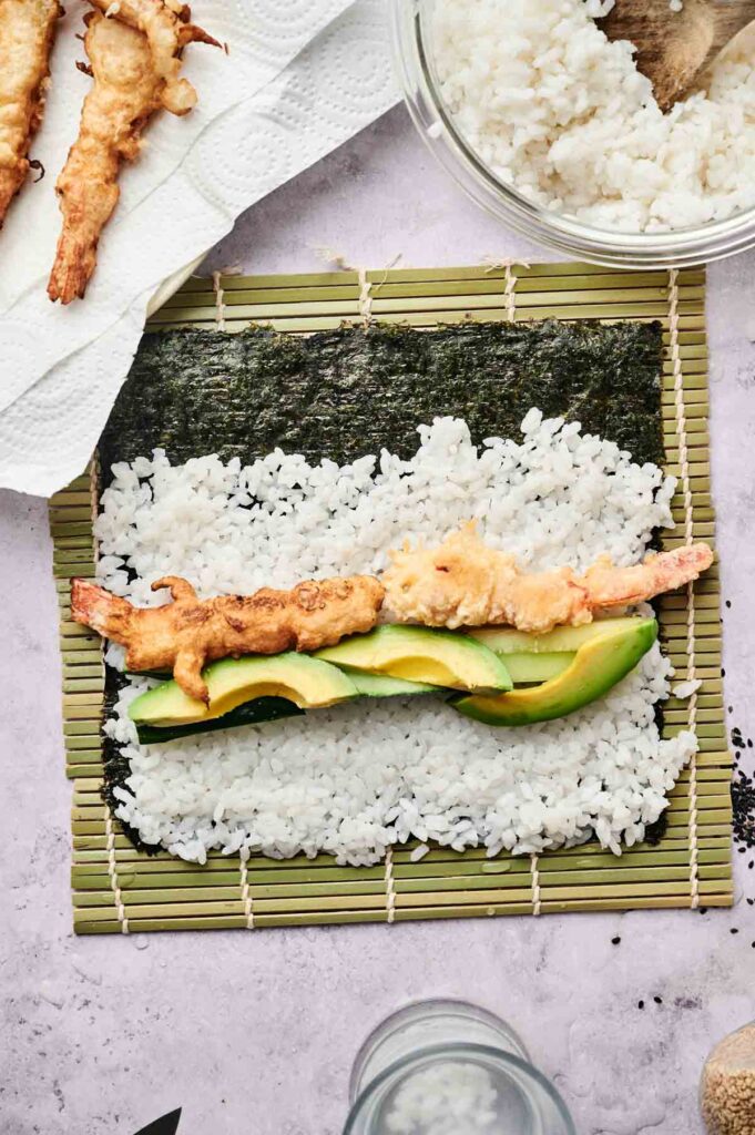 A sushi mat with a sheet of nori topped with sushi rice, sliced avocado, and tempura shrimp, ready to be rolled into a classic Shrimp Tempura Roll.