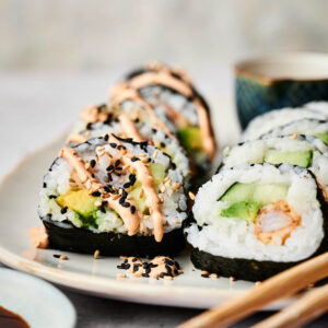 A plate of Shrimp Tempura Roll sushi filled with avocado and cucumber, topped with spicy mayo and black sesame seeds, served with chopsticks and a small bowl of soy sauce on the side.
