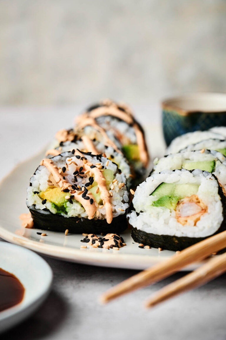 A plate of Shrimp Tempura Roll sushi filled with avocado and cucumber, topped with spicy mayo and black sesame seeds, served with chopsticks and a small bowl of soy sauce on the side.