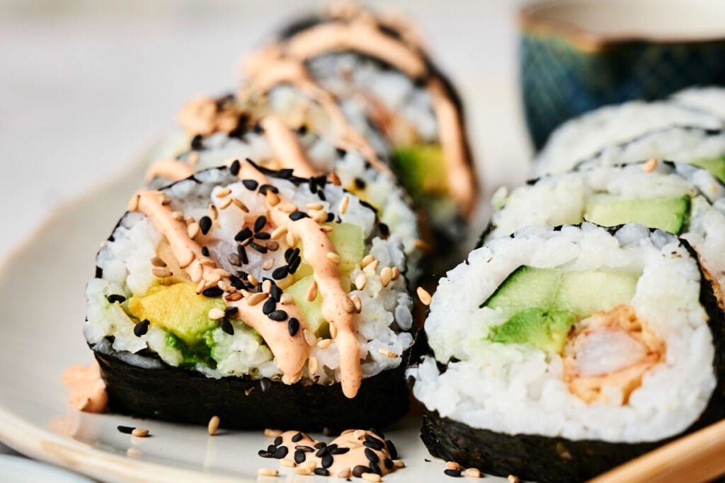 Close-up of a Shrimp Tempura Roll with rice, avocado, and shrimp, topped with sesame seeds and a drizzle of creamy sauce, beautifully arranged on a plate.