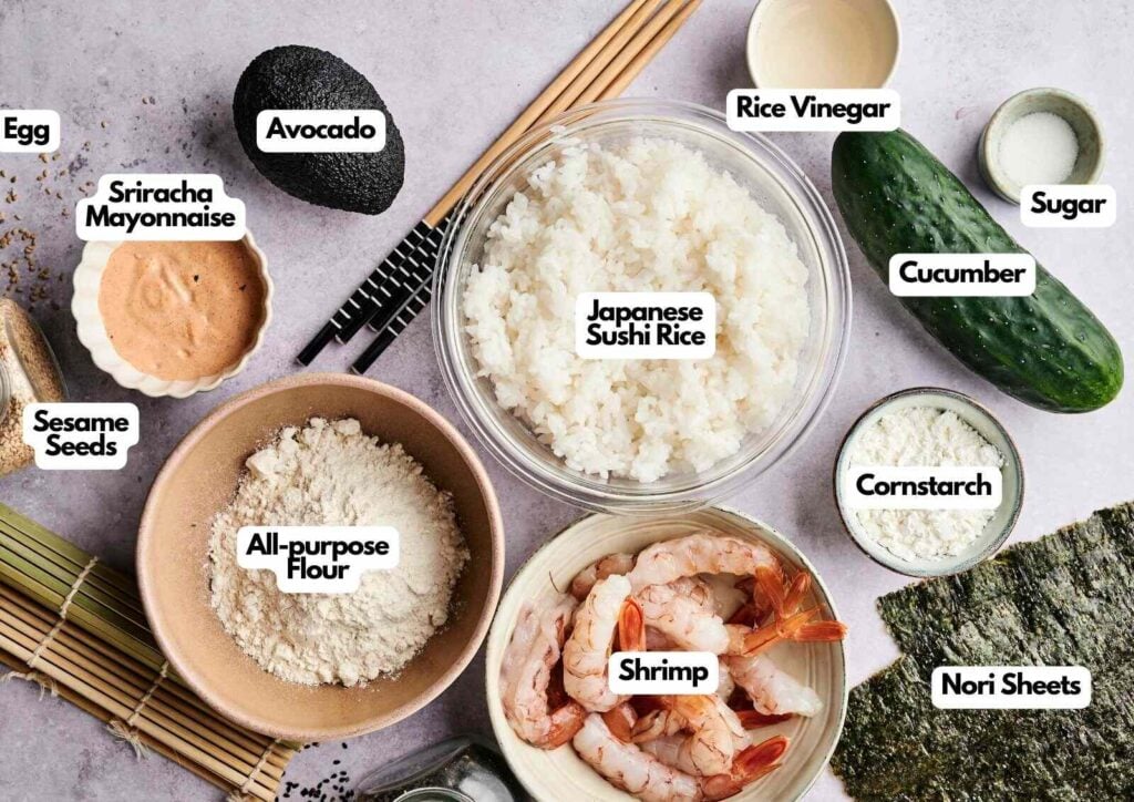 Ingredients for a Shrimp Tempura Roll are arranged on a surface, including shrimp, avocado, cucumber, sushi rice, nori sheets, egg, flour, sugar, rice vinegar, sriracha mayo, cornstarch, and sesame seeds.