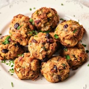 A white plate holds a pile of stuffing balls garnished with chopped herbs, featuring visible pieces of vegetables and cranberries for a festive touch.