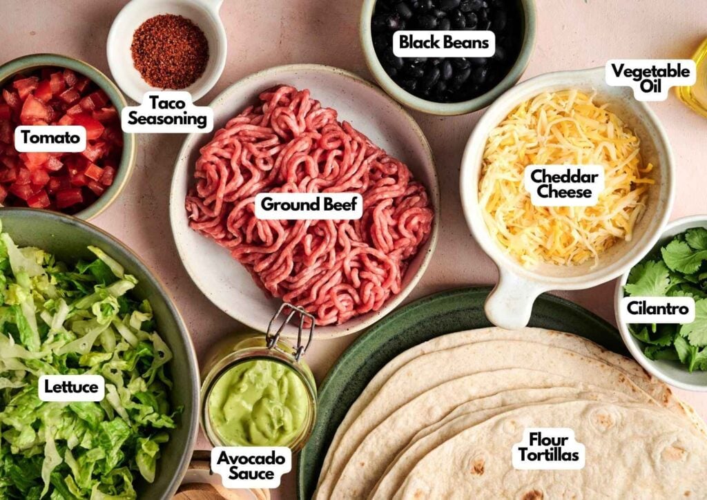 A flat lay of taco ingredients, perfect for making a Taco Salad, includes ground beef, flour tortillas, cheddar cheese, lettuce, tomato, avocado sauce, black beans, cilantro, vegetable oil, and taco seasoning.